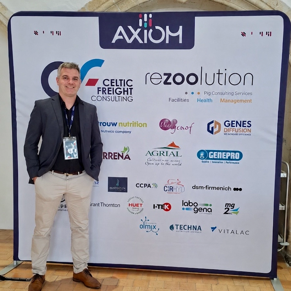 Homem sorrindo de terno cinza e calça clara em frente ao painel de patrocinadores do AXIOM International Summit em Rennes, França, destacando o logotipo da GENEPRO entre as marcas parceiras de tecnologia em reprodução suína.