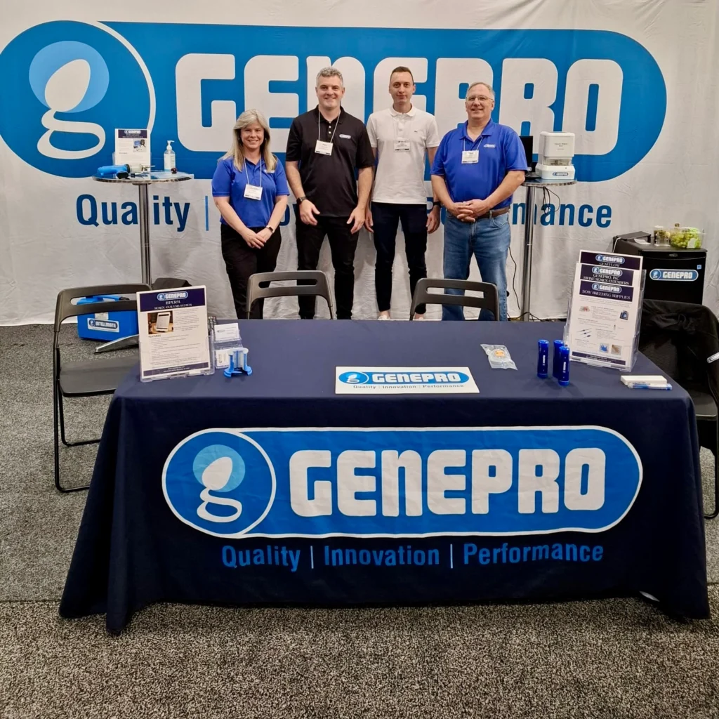 Quatro integrantes da equipe GENEPRO sorrindo atrás de uma mesa com toalha azul com o logotipo da marca na World Pork Expo 2026. O estande exibe fôlderes informativos, soluções para reprodução suína e um grande painel ao fundo com o slogan "Quality, Innovation, Performance".