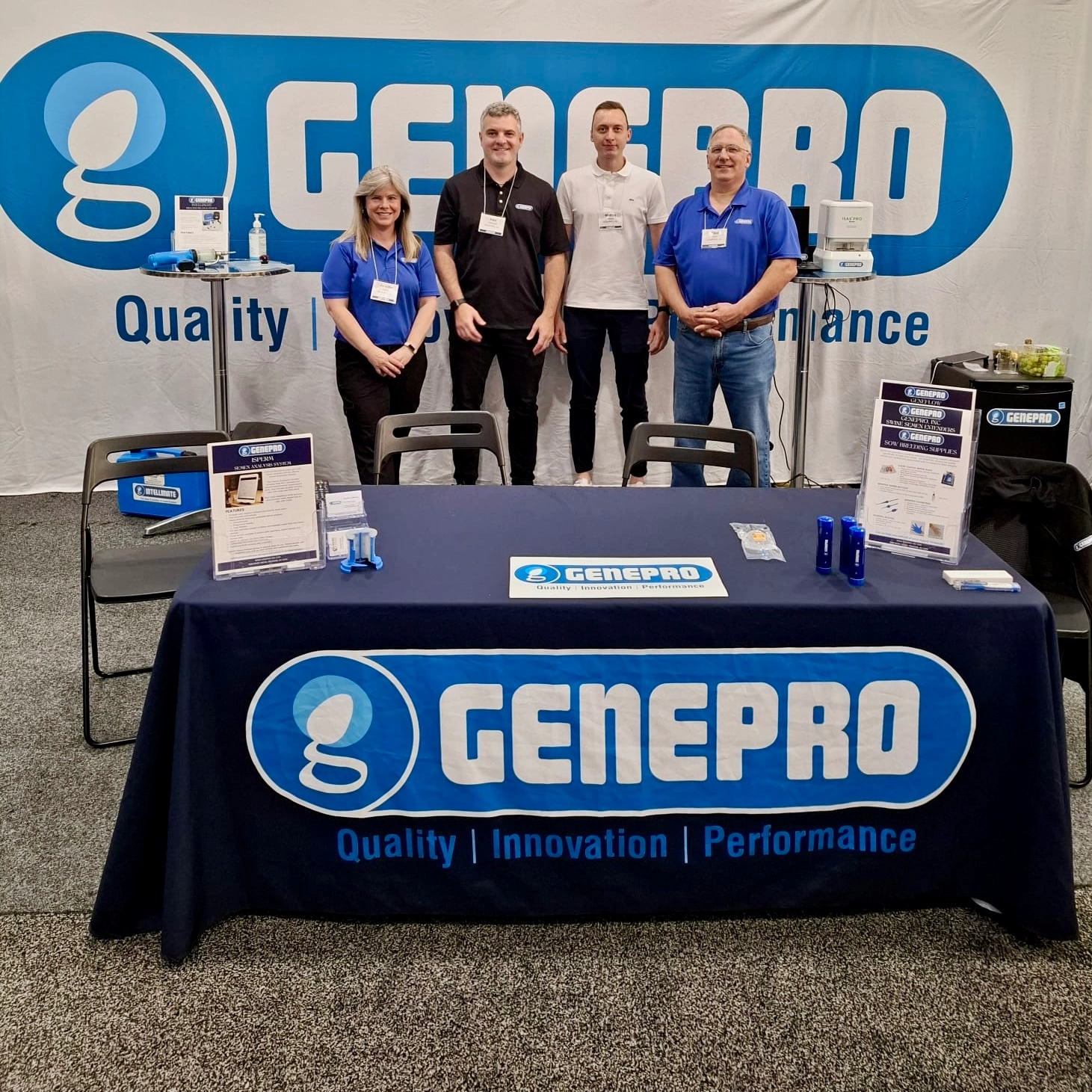 Quatro integrantes da equipe GENEPRO sorrindo atrás de uma mesa com toalha azul com o logotipo da marca na World Pork Expo 2026. O estande exibe fôlderes informativos, soluções para reprodução suína e um grande painel ao fundo com o slogan "Quality, Innovation, Performance".
